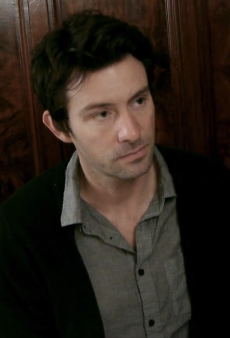 一个有黑色短发的男人，穿着灰色扣子衬衫和黑色开衫。