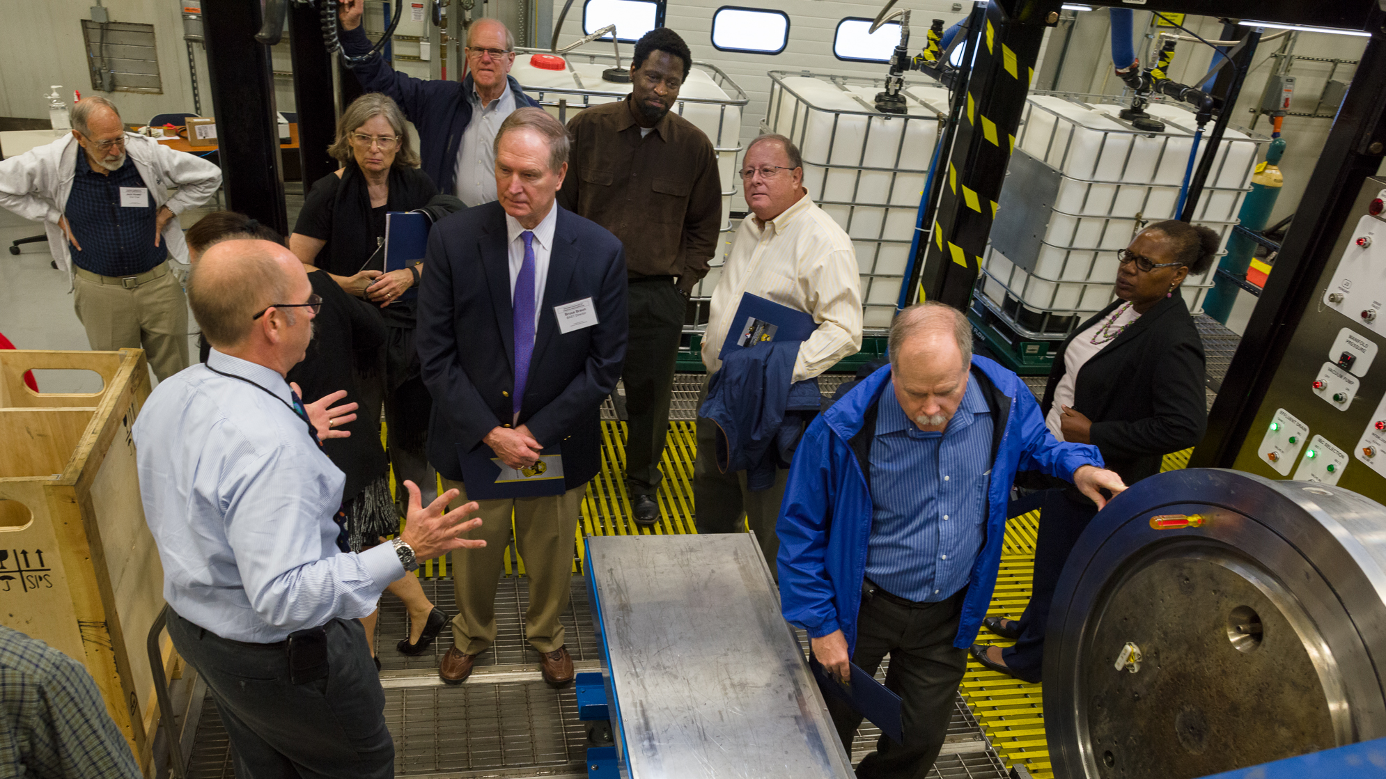 一群衣着讲究的人在车间里聆听一名男子讲话。